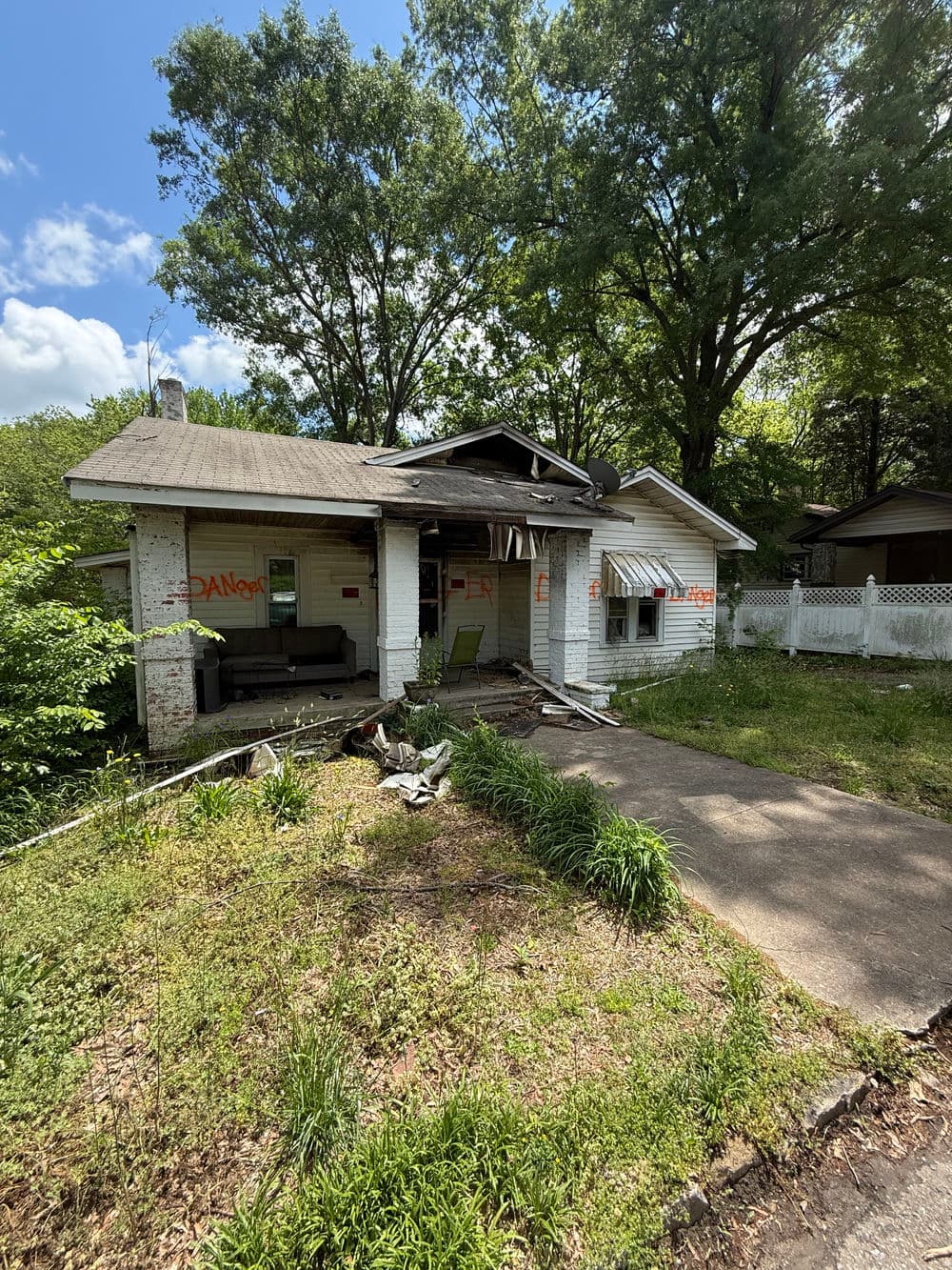 Deteriorating abandoned house with overgrown grass and visible damage in the front yard.