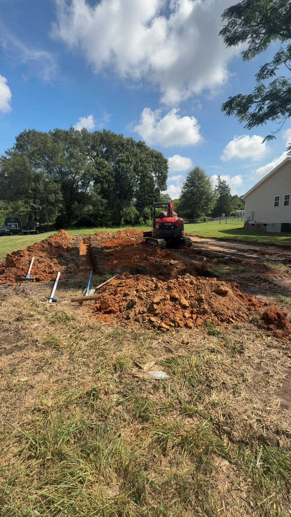 Excavation site with red machinery, dirt piles, and plumbing pipes under a clear blue sky.