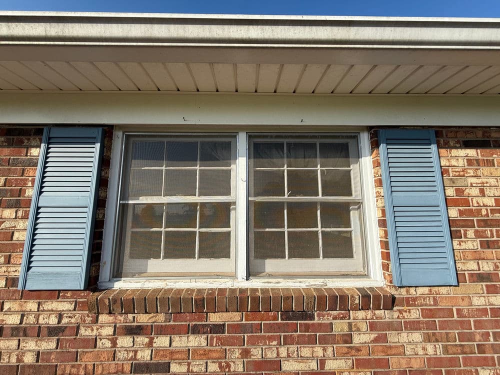 Brick house exterior featuring a window with blue shutters and a clear blue sky.