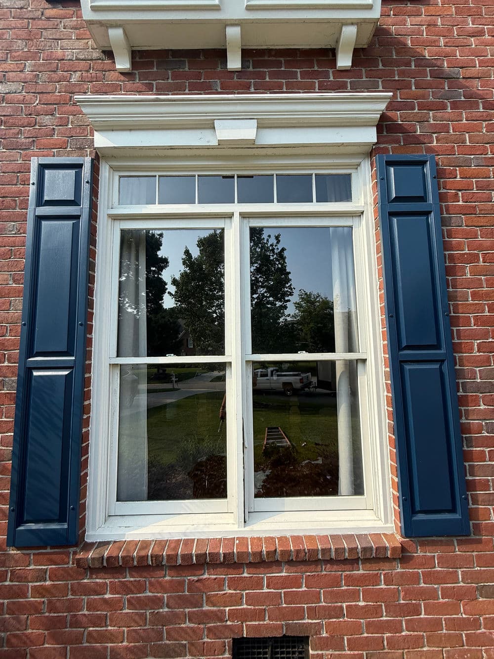 Classic brick house window with blue shutters and reflections of surrounding greenery.