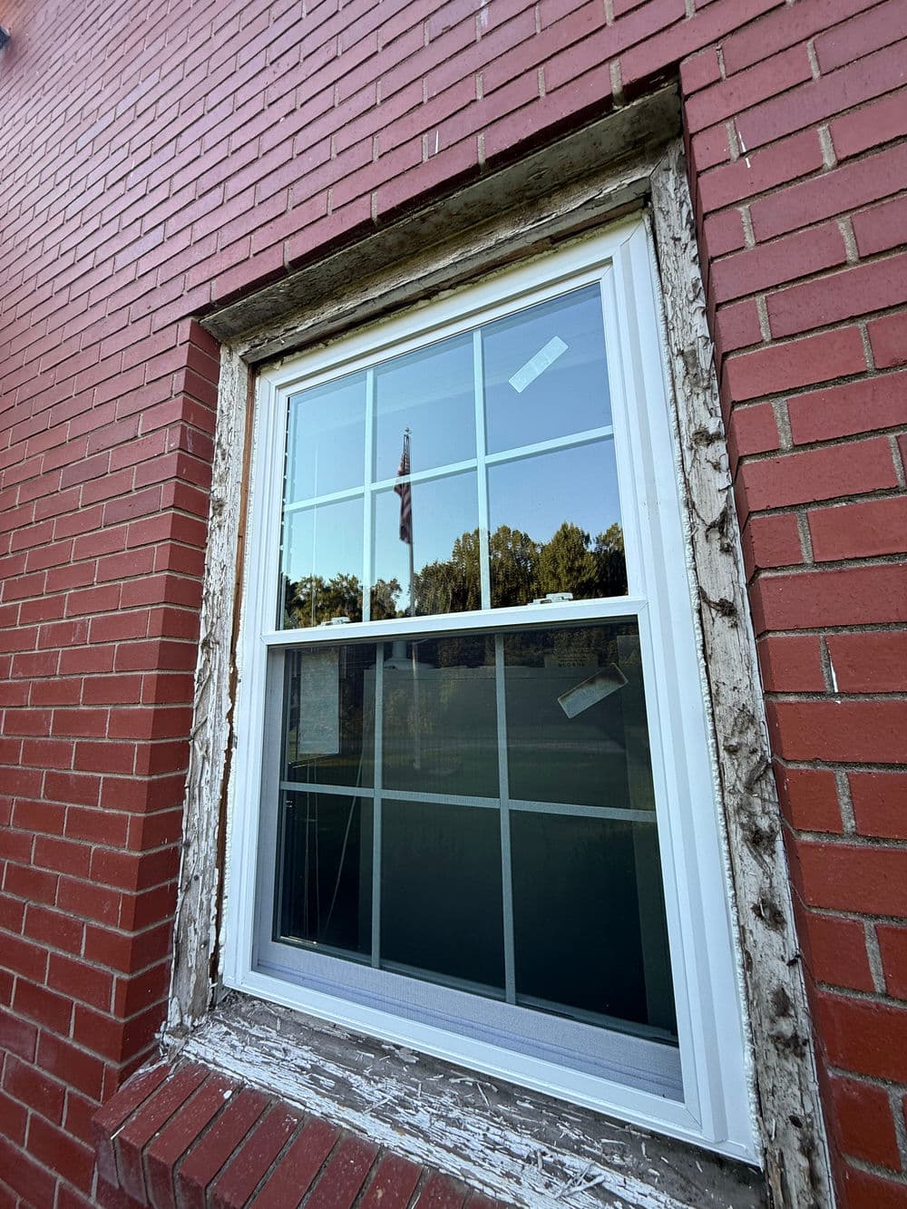 Window with peeling trim in red brick wall, reflecting trees and a flag in the glass.