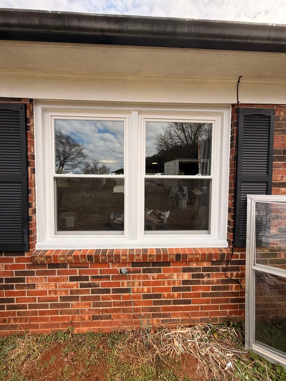 Newly installed double window with white frames and black shutters on a red brick house exterior.