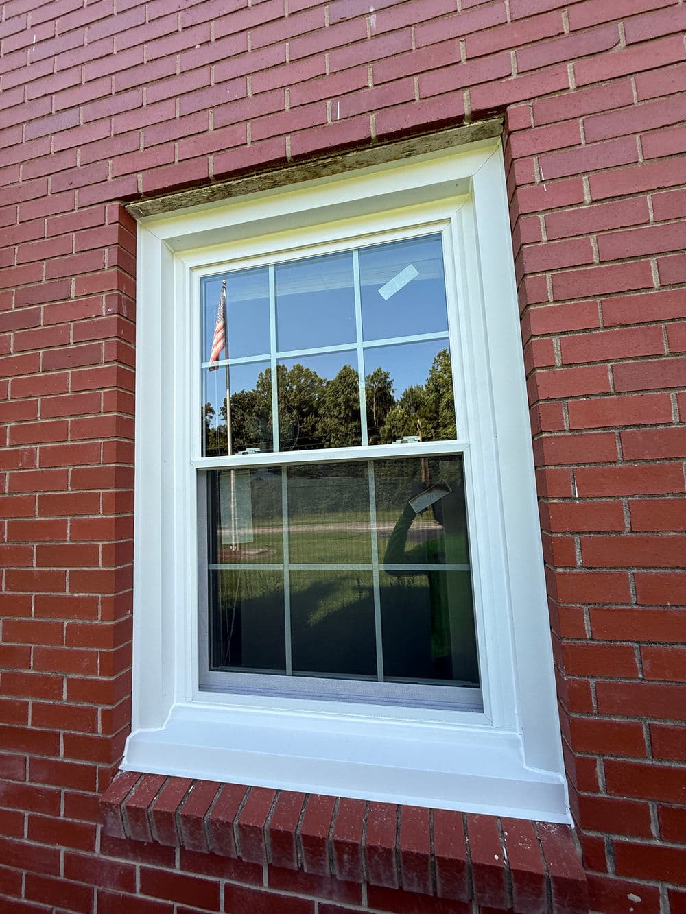 White window frame on a brick wall reflecting trees and an American flag outside.