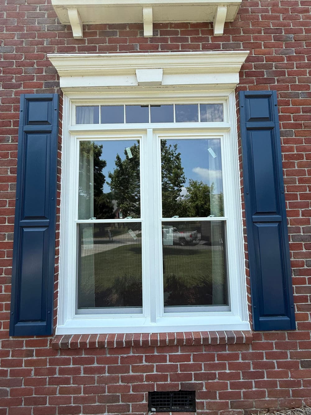 Traditional brick home window with blue shutters and clear glass reflecting greenery outside.